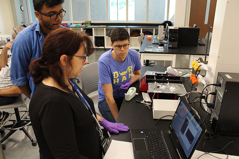 Abhik Manna (left) and Nathan Whitman (right) listen and watch as Maggie Witek discusses data collection with a micro Coulter Counter device