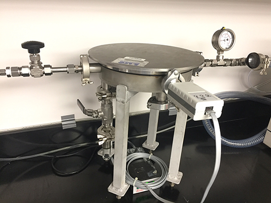 Metallic CVD chamber with various valves, gauges, and connectors on a lab bench.
