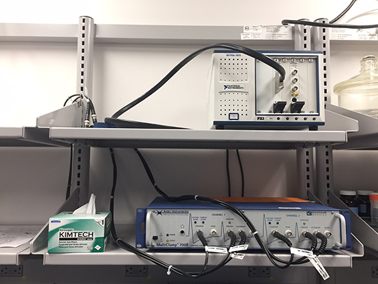A patch clamp setup with amplifiers and multiple channel controllers on laboratory shelves.