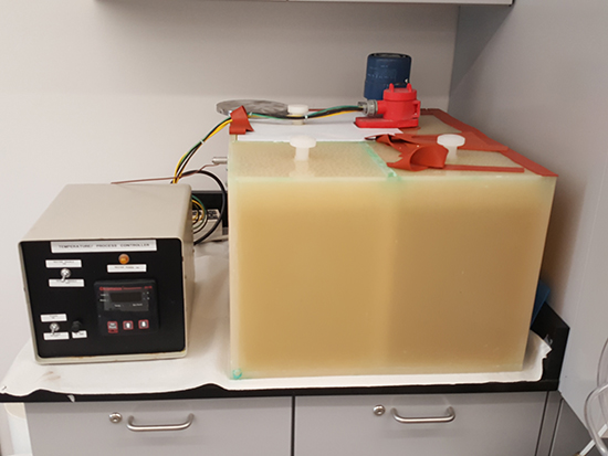 A lab setup featuring a plating apparatus with a large transparent container filled with a yellowish solution, and an adjacent temperature controller unit.