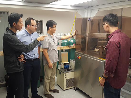 Alt text: "Daniel, Yijie, Xiaoxiao, and Daewon discussing around a laboratory equipment, with Xiaoxiao pointing out details using a yellow rod.