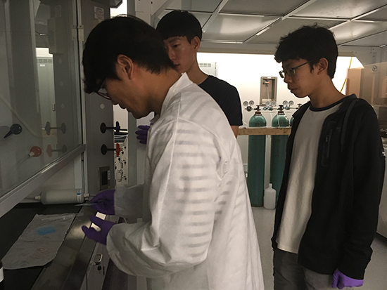 Junseo, Daewon, and Sunggun working at a spin-coater station, with Junseo handling an object while the others observe.
