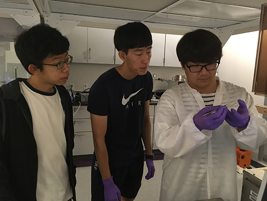Junseo, Daewon, and Sunggun observing an item that Junseo is holding, while in a laboratory setting with equipment in the background.