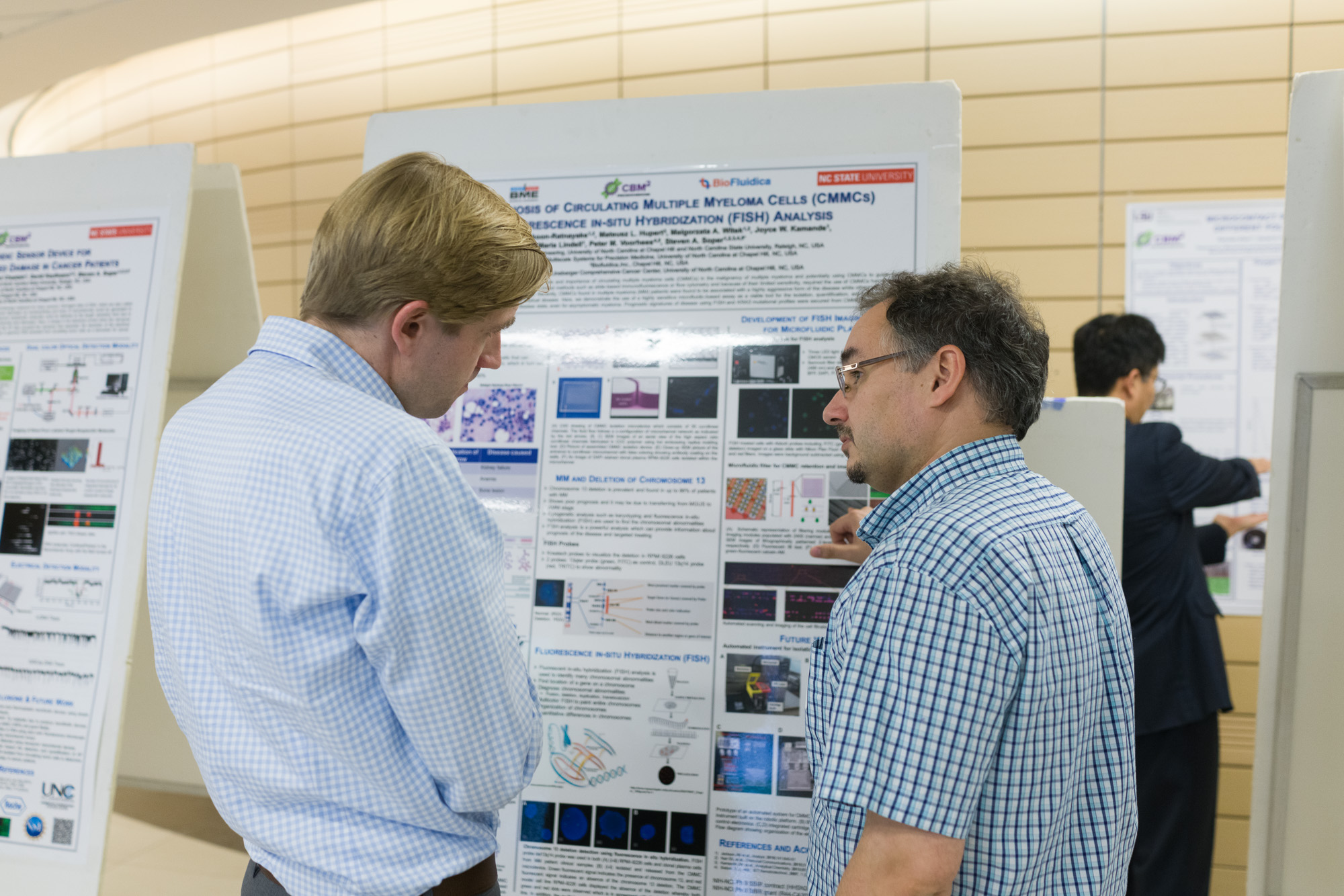Two men discussing a scientific poster about 'Circulating Multiple Myeloma Cells' and 'FISH Analysis', with other posters in the background.
