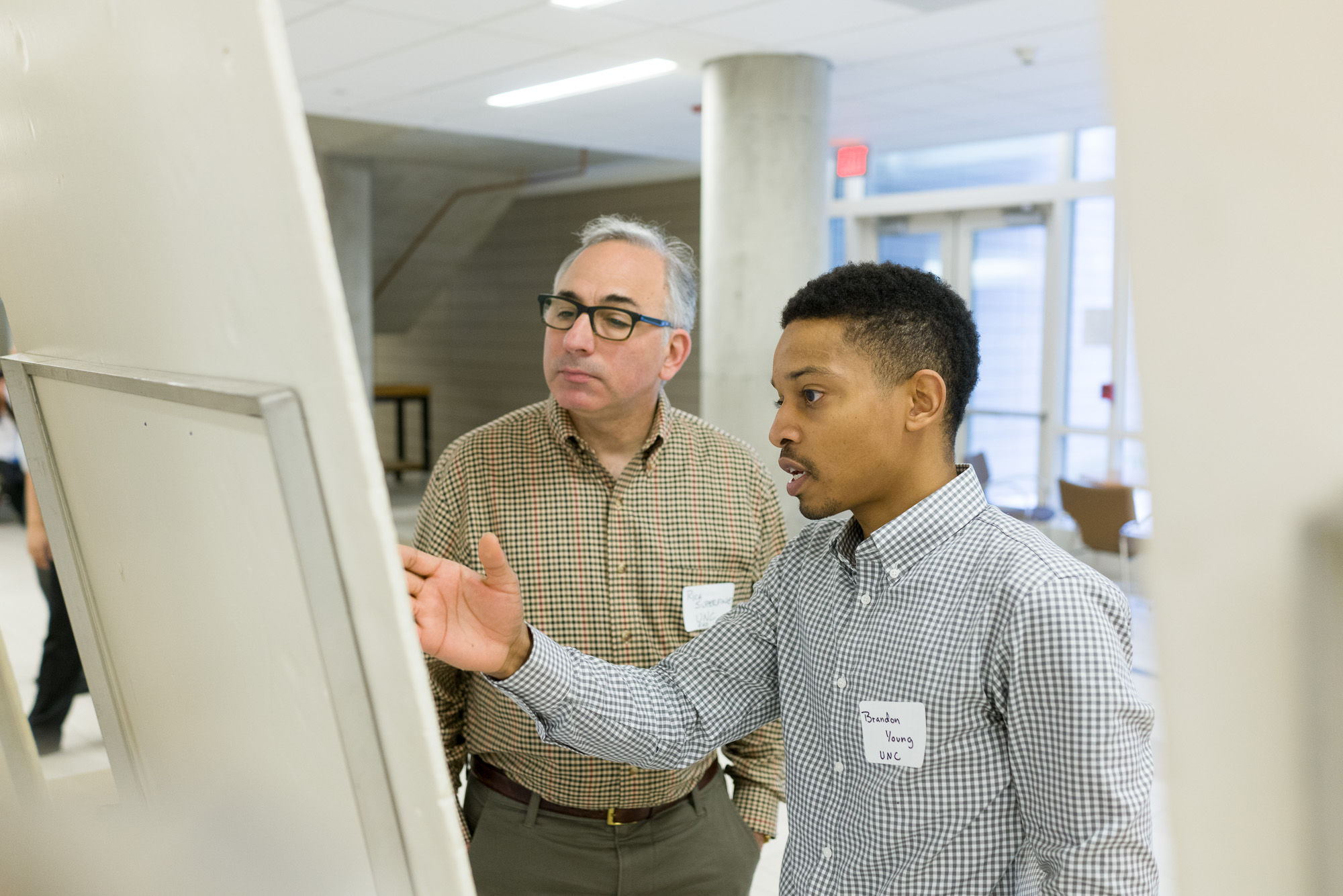 Two men discussing in front of a research poster; one wears a name tag labeled 'Brandon Young - UNC'.