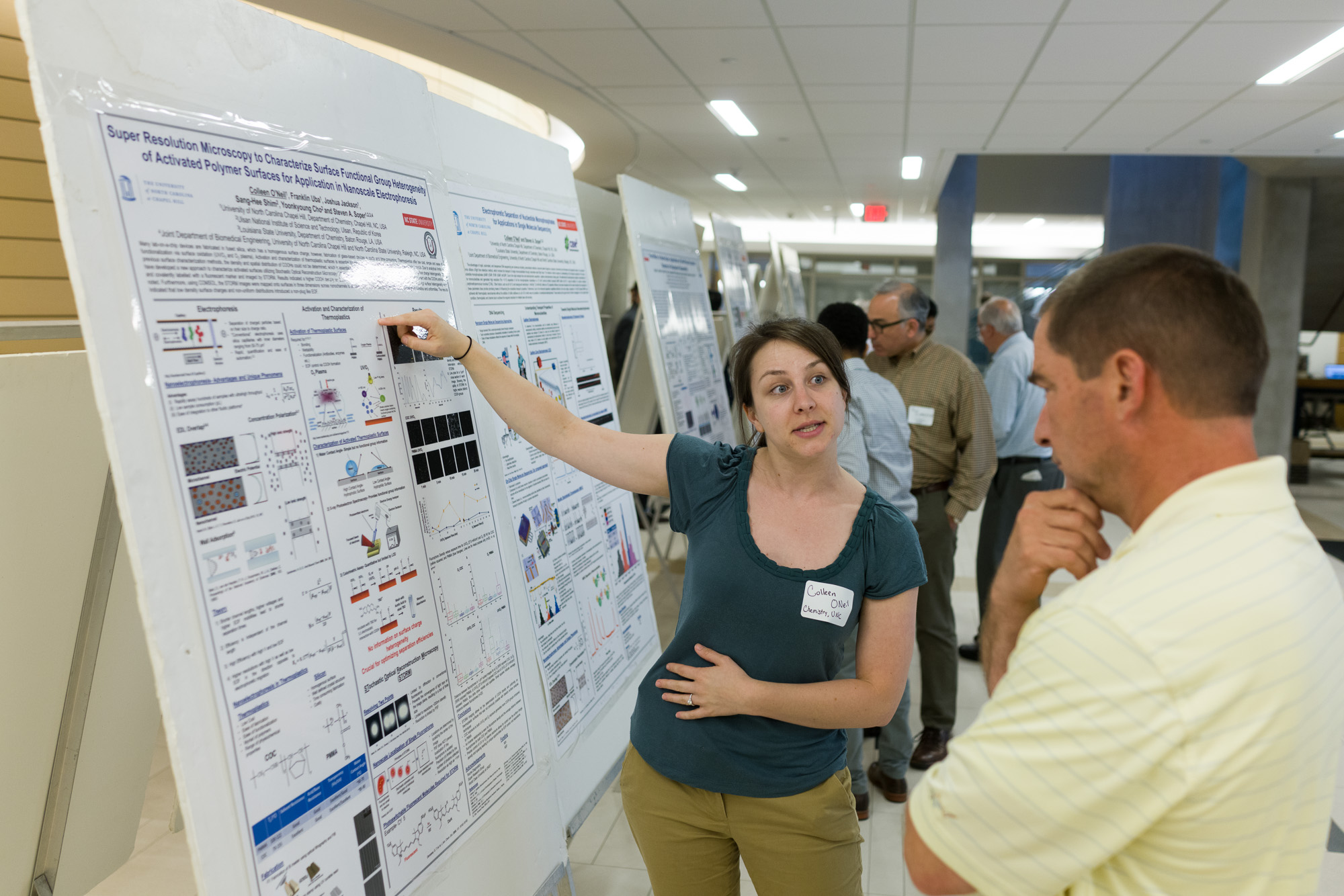 A woman with a nametag labeled 'Colleen' presents scientific data on a large poster, pointing to a section, while discussing with a man in a striped shirt. The backdrop features other attendees observing similar posters.