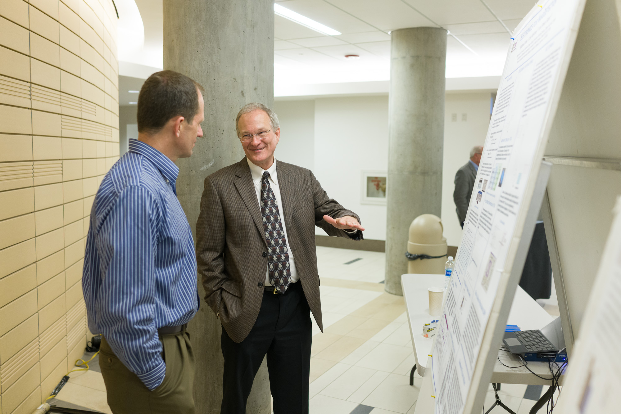 Two men converse near a scientific poster at a conference, with one gesturing towards something.