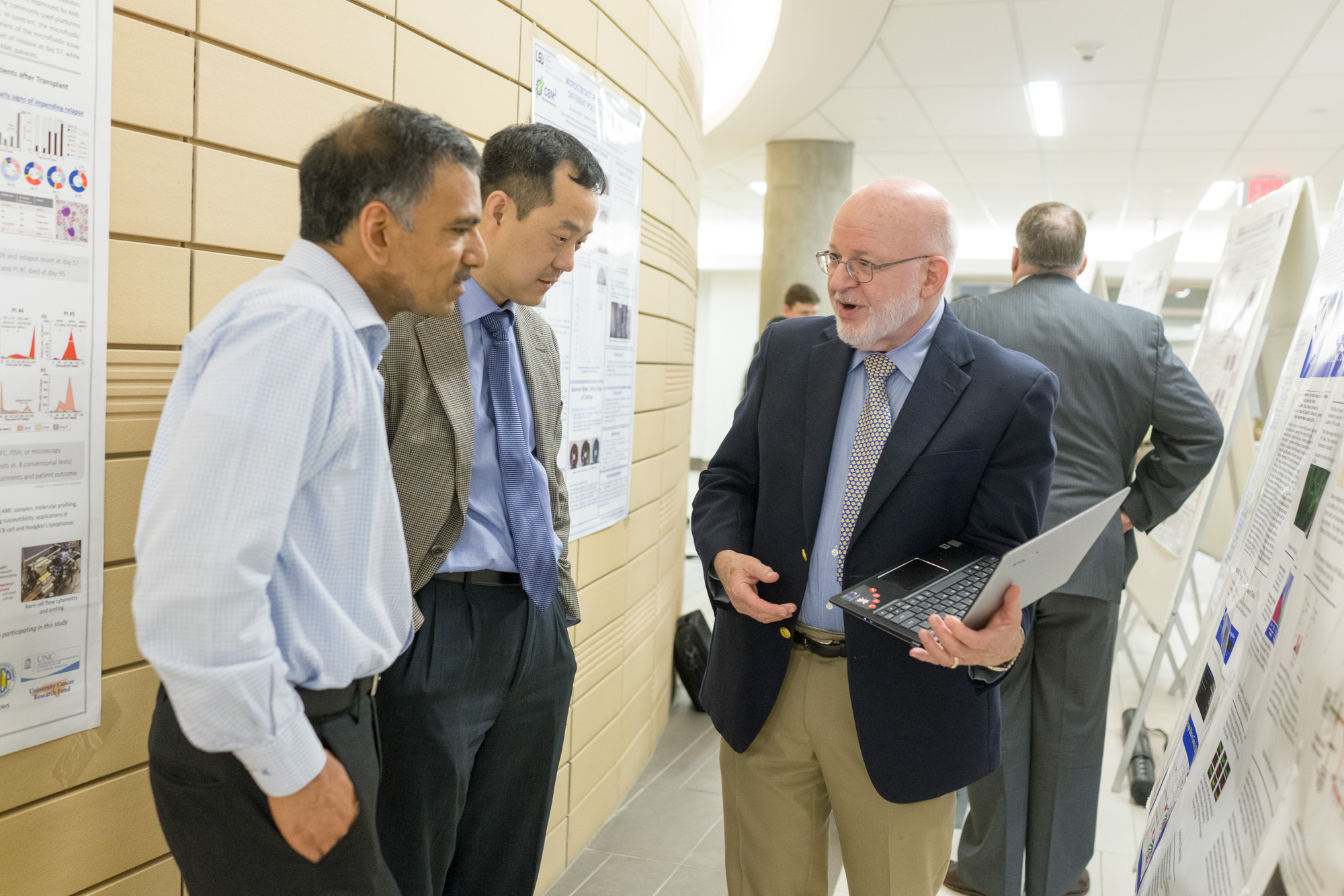 Three professionals discuss scientific posters in a hallway. One gentleman with a laptop shares insights with two other men.