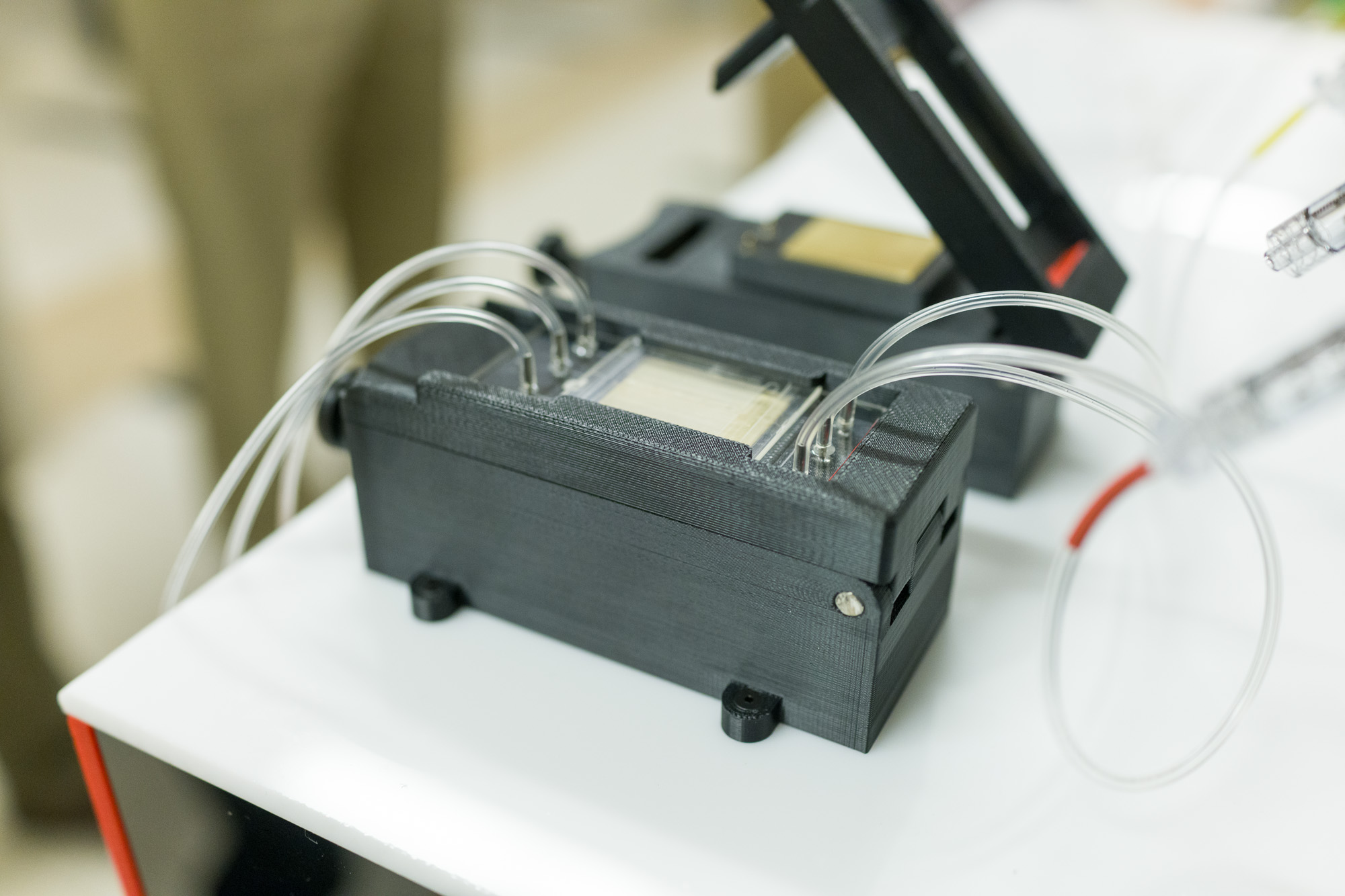 A close-up of a black, 3D-printed device with transparent tubes attached, resting on a white surface with a red edge.