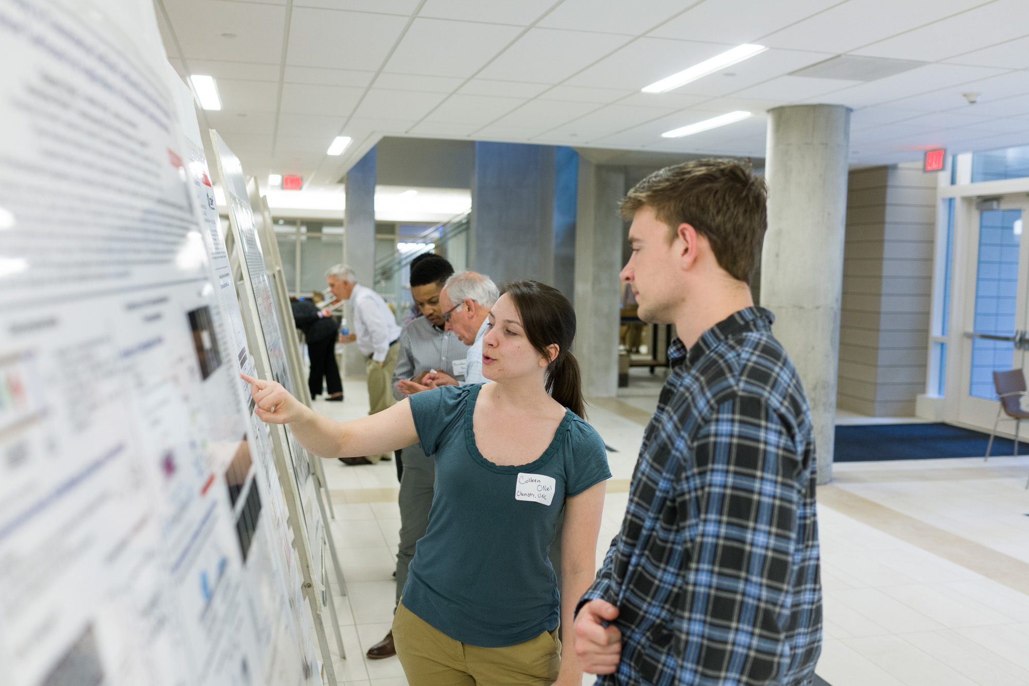 A woman with a name tag 'Caitlin Oke, University' points at a poster board, discussing its contents with a man beside her. Other attendees are seen examining posters in a well-lit indoor venue.