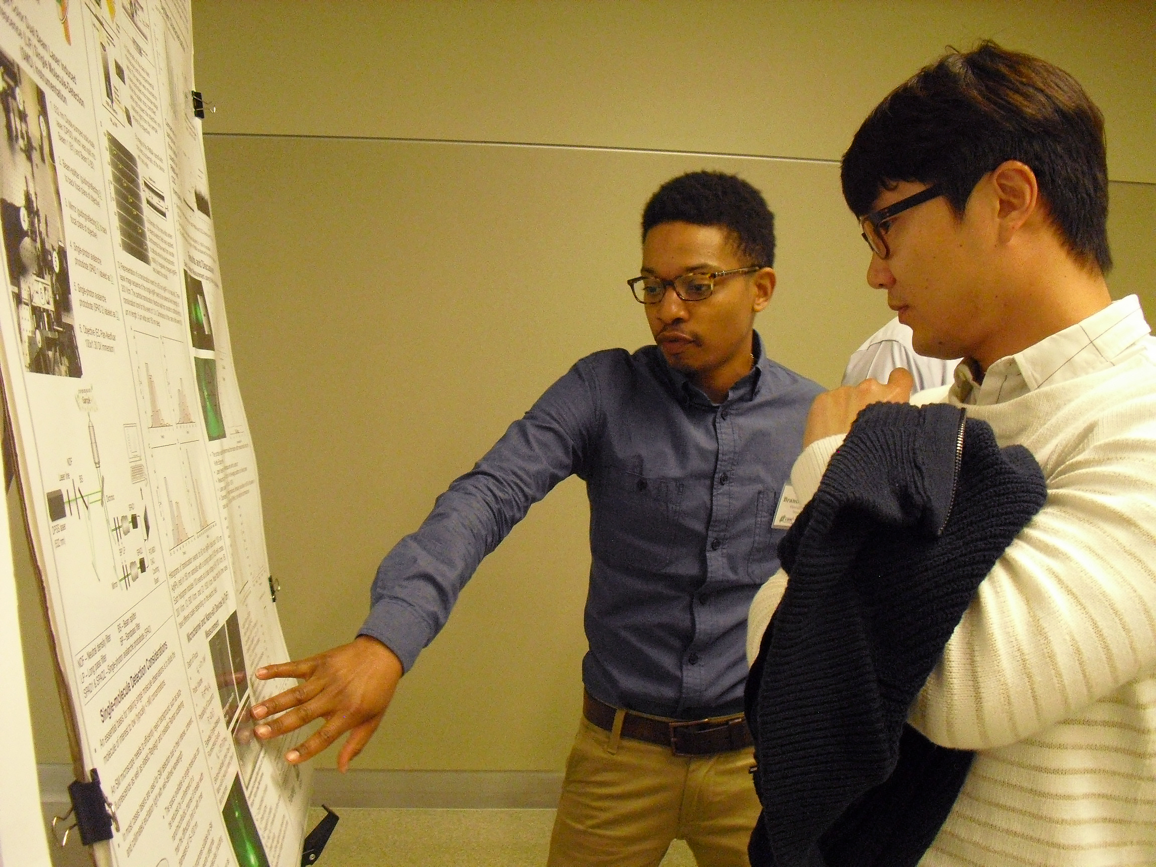 Two individuals standing in front of a single research poster, one actively pointing to elements on it.