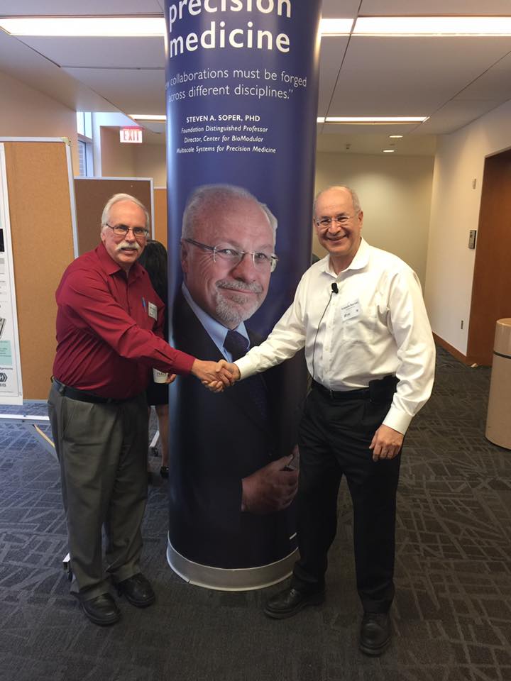 Two individuals shaking hands in front of a pillar, with a prominent poster featuring Dr. Soper in the background.