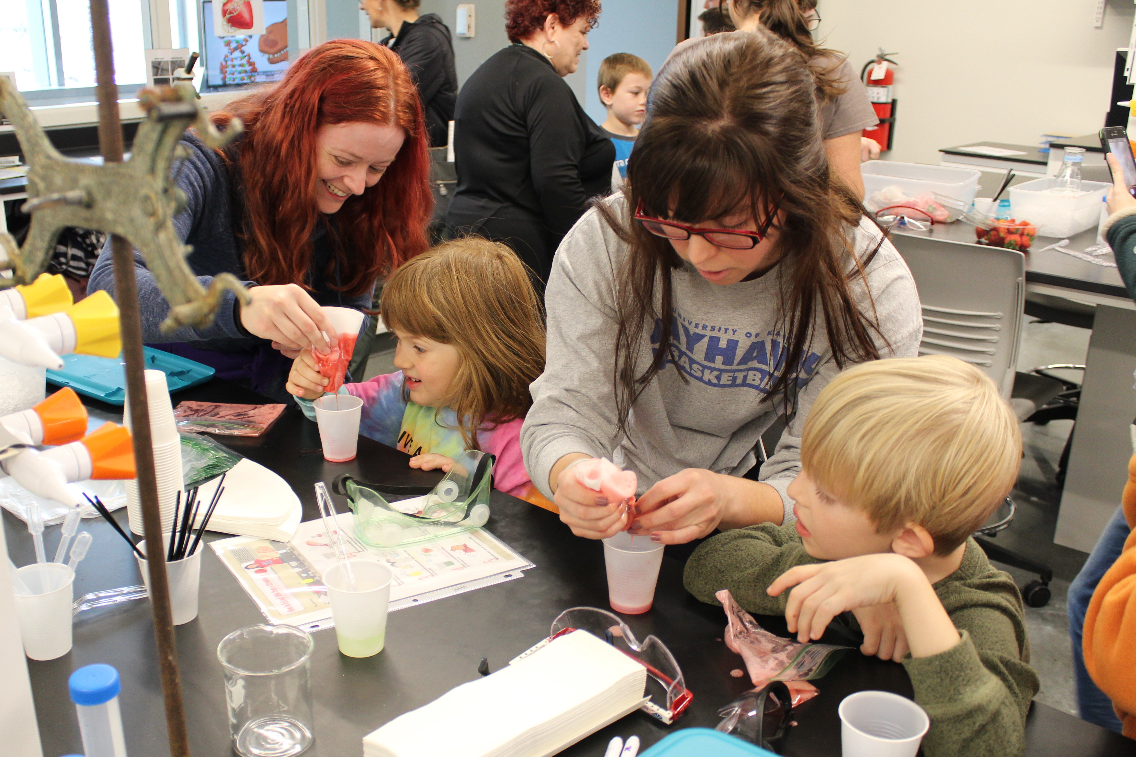Two adults assisting two children with handling cups filled with red colored liquid.