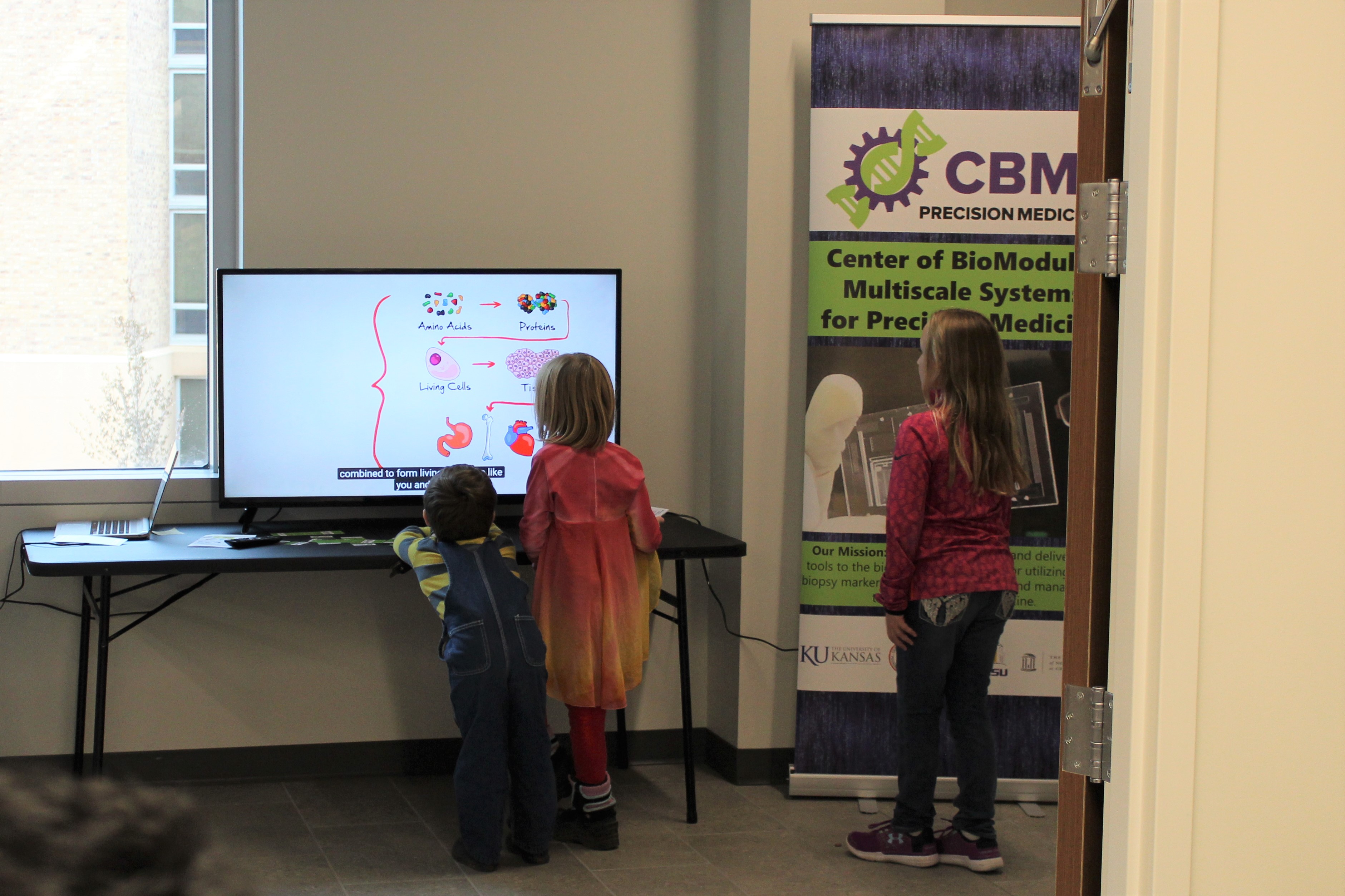 Three children watching chemistry demonstrations on a TV screen.