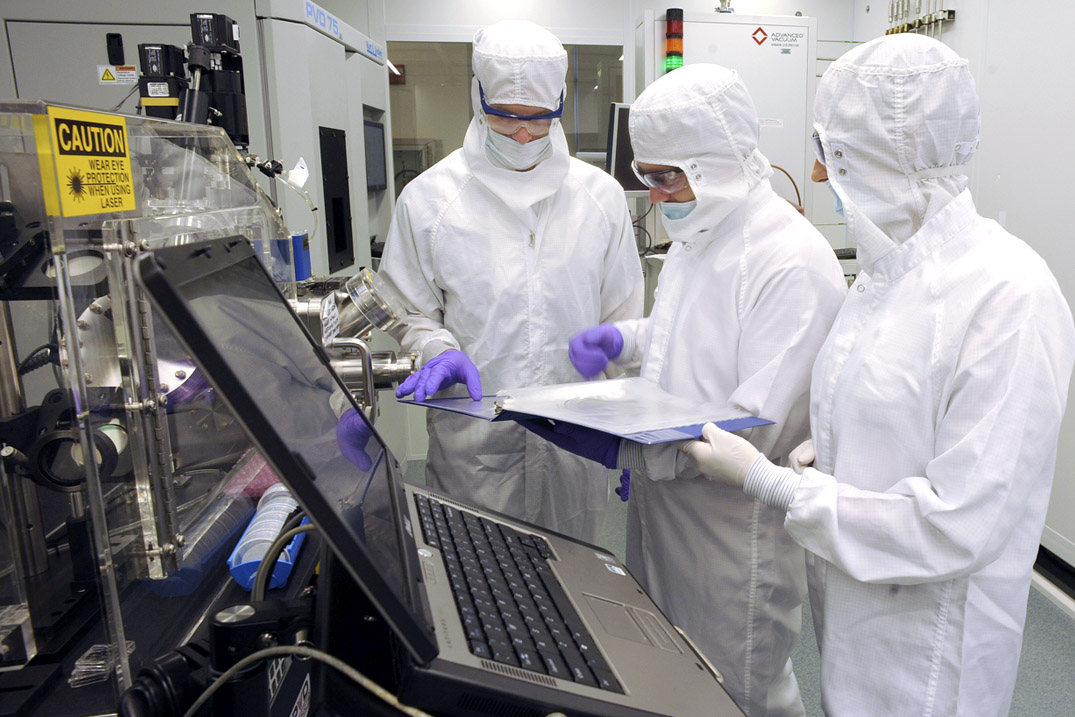 Three individuals in cleanroom suits examining a document near machinery in a lab.