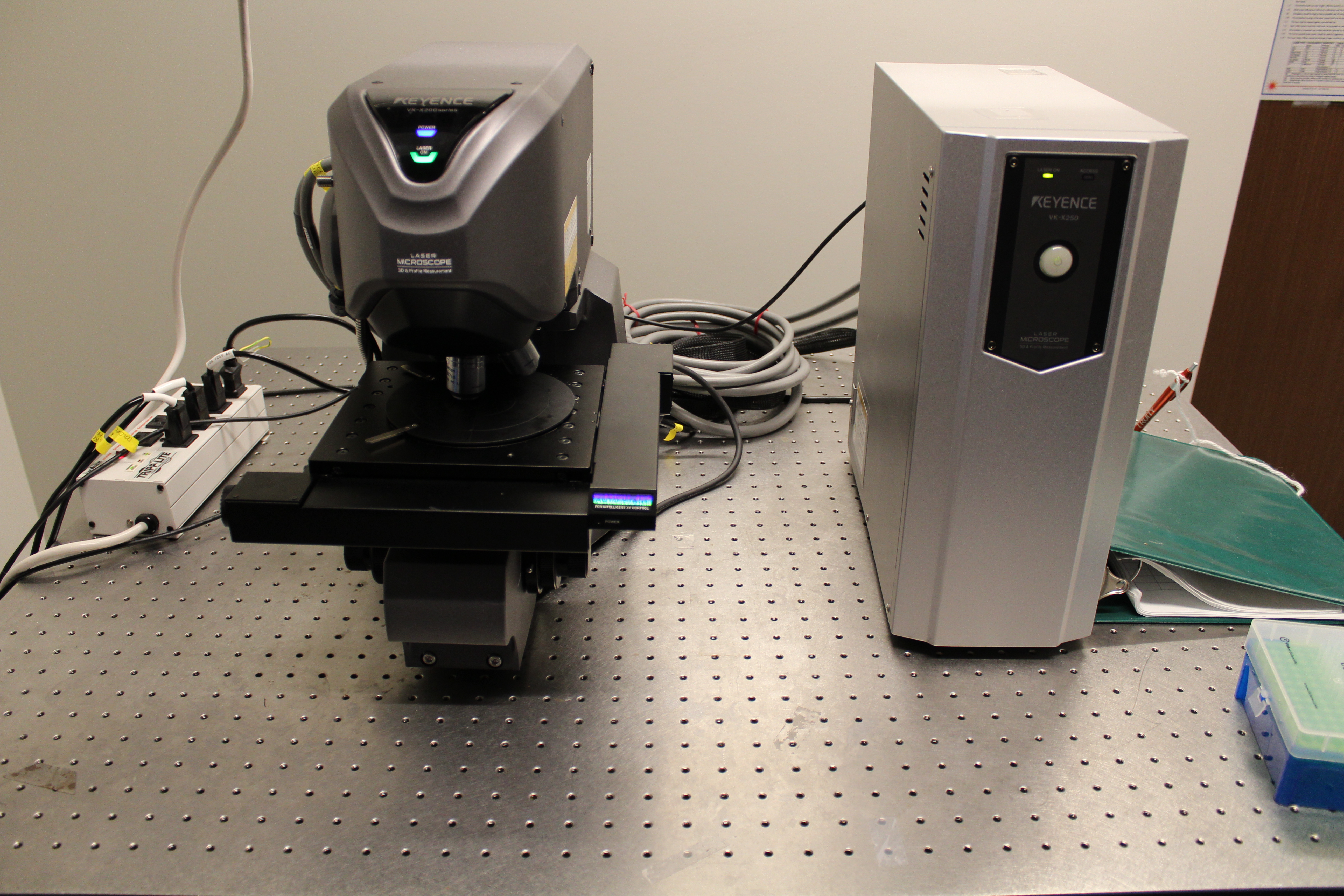 Laboratory setup with a precision microscope and electronic unit on a metal table, with cables and a face mask nearby.